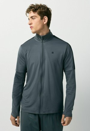 Young man wearing a dark gray zip-up athletic jacket with long sleeves, standing against a plain light background, looking left.