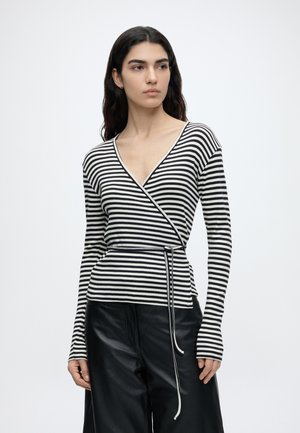 Femme aux longs cheveux foncés portant un haut cache-cœur rayé noir et blanc avec ceinture à nouer et un pantalon noir à jambes larges, debout devant un fond uni.