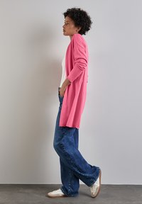 Rosa lång kofta i mjukt material, bärs över en vit topp, matchad med blå vida jeans och ljusa sneakers.