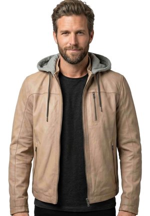 Mann mit Bart und gestyltem Haar, der eine beige Lederjacke mit grauer Kapuze und schwarzem T-Shirt trägt, blickt nach vorne vor weißem Hintergrund.