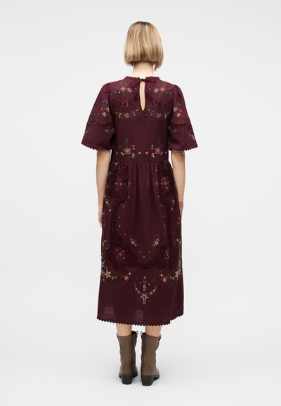 Γυναίκα με κοντά ξανθά μαλλιά που φοράει μίντι φόρεμα βουργουνδί με φουσκωτά μανίκια, κεντήματα με λουλούδια και καφέ μποτάκια αστραγάλου, εμφανιζόμενη από πίσω.