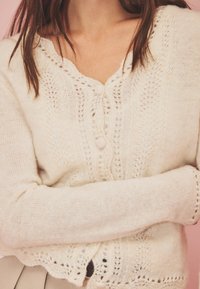 Cárdigan beige claro con un delicado patrón de encaje, escote redondeado y cierre de botones. Textura suave, mangas largas y dobladillo con puntilla.