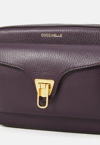 Borsa in pelle di colore viola scuro con una finitura testurizzata. Presenta una chiusura in metallo dorato e un logo impresso sul davanti. Bordo liscio.