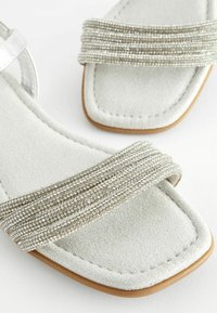 Vlakke sandalen met een lichtgrijze suède voetbed en decoratieve zilveren kralenbandjes. Vierkante neusontwerp met een bruine rubberen zool.