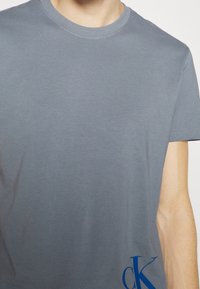 Grå bomullströja med rund hals, korta ärmar och en blå logotyp i det nedre högra hörnet. Slät textur med minimala designdetaljer.