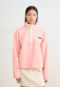Pull en polaire rose clair avec un col haut, des boutons crème et une petite poche zippée. Présente un empiècement de logo à l'avant.