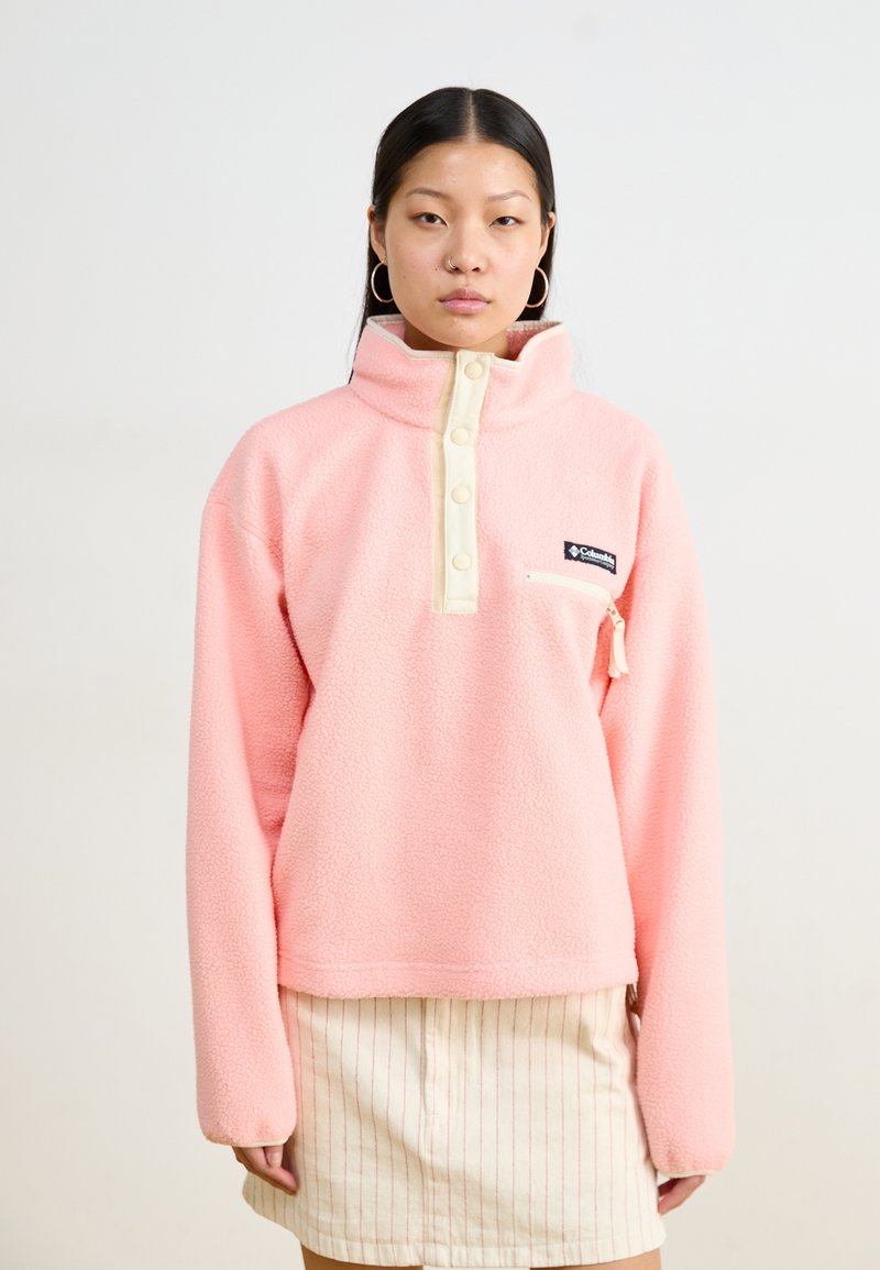 Pull en polaire rose clair avec un col haut, des boutons crème et une petite poche zippée. Présente un empiècement de logo à l'avant.