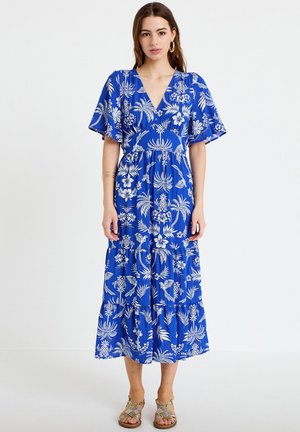 Kvinde iført en blå blomster-printet midikjole med flagrende ærmer og guld sandaler, stående mod en ensfarvet hvid baggrund.