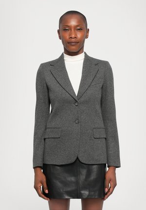 Woman wearing a grey herringbone blazer over a white turtleneck and a black leather skirt, standing against a plain white background.