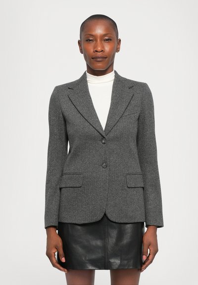 Donna che indossa un blazer grigio a spina di pesce sopra un dolcevita bianco e una gonna di pelle nera, in piedi contro uno sfondo bianco semplice.
