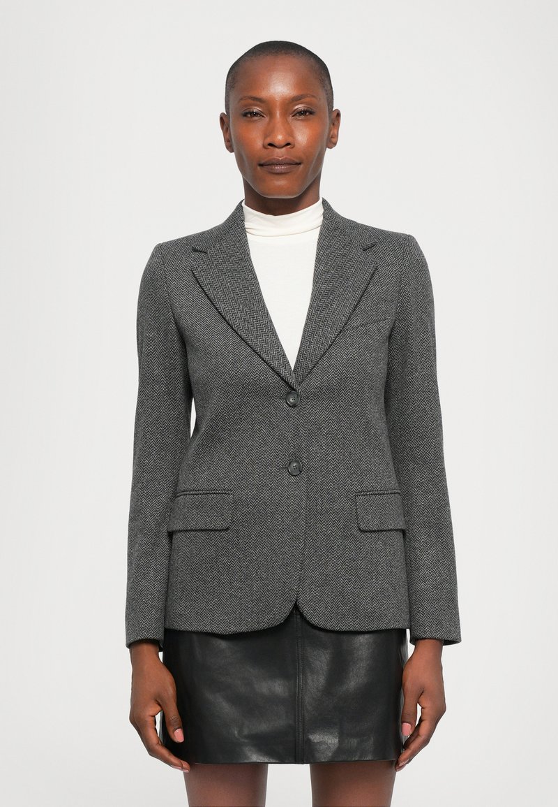 Vrouw in een grijze herringbone blazer over een witte turtleneck en een zwarte leren rok, staand tegen een effen witte achtergrond.