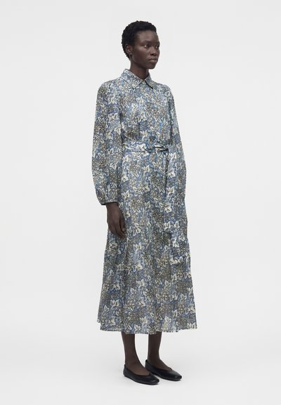 Γυναίκα που στέκεται, φοράει ένα μακρυμάνικο, ανοιχτό μπλε φόρεμα με floral σχέδιο μέχρι τον αστράγαλο, με γιακά και μαύρα επίπεδα παπούτσια, σε απλό λευκό φόντο.