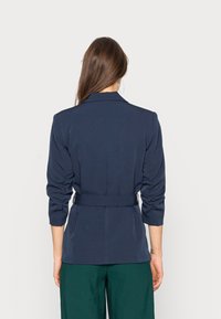Blazer bleu marine à coupe ajustée, manches trois-quarts froncées et ceinture assortie à la taille. Tissu lisse, design minimaliste.