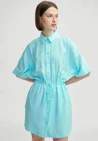 Robe courte à manches courtes bleu clair avec un devant boutonné, une taille à cordon et une silhouette décontractée. Fabriquée dans un tissu lisse et respirant.