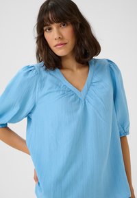 Light blue blouse with a V-neck, short puffed sleeves, and textured fabric. Features subtle pleats at the neckline and relaxed fit.