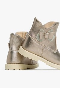 Metallic gold Ankle Boots mit strukturiertem Finish, ausgestattet mit einer hellen Gummisohle und einem Stoffbogenakzent auf der Rückseite.