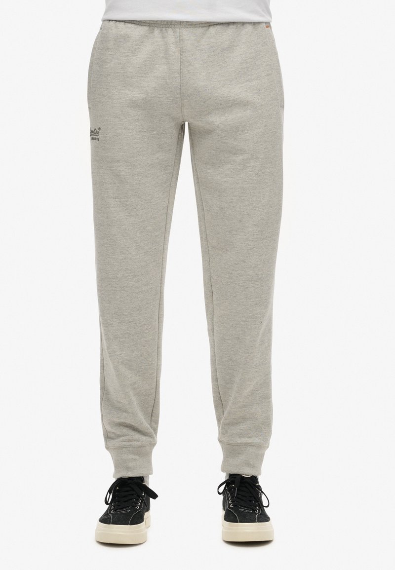 Un pantalon de survêtement gris en tissu doux, avec une taille élastique, des jambes fuselées et une marque discrète près de la hanche, associé à des baskets noires.