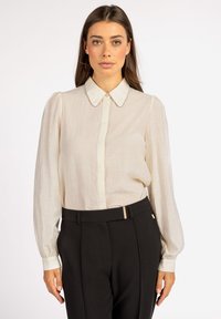 Cream blouse with a classic collar, puffed sleeves, and button-down front; paired with black tailored pants featuring a gold accent.