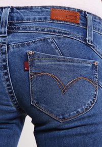 Vaqueros de mezclilla azules con un parche de cuero marrón en la cintura y un distintivo bordado en forma de corazón en el bolsillo trasero.