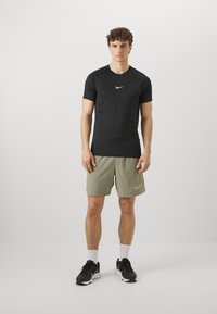 Maglietta Nike nera a maniche corte con logo bianco, abbinata a pantaloni corti verde chiaro e scarpe da ginnastica nere. Design semplice e funzionale.