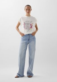 White graphic t-shirt with colorful design, paired with light blue wide-leg jeans, featuring a casual fit and pockets. Sandals complete the look.