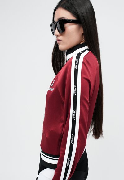 Červená bunda na zip se černými a bílými bočními pruhy, s grafikou "OFF-White". Model má na sobě černé obdélníkové sluneční brýle. Hladká textura.