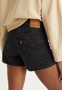 Persona que lleva puestos unos pantalones cortos de mezclilla negra de Levi's y una camisa de manga larga beige, mostrando la vista trasera y la etiqueta de la marca en los pantalones cortos.
