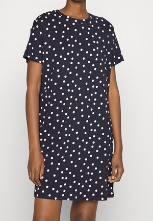 Personne portant une robe bleu marine manches courtes à pois blancs, debout devant un fond clair uni.