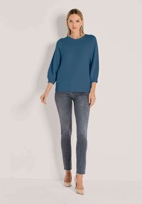 Frau trägt einen blauen Strickpullover mit Dreiviertelärmeln, graue Skinny-Jeans mit fransigen Säumen, beige High Heels und hängende Ohrringe.