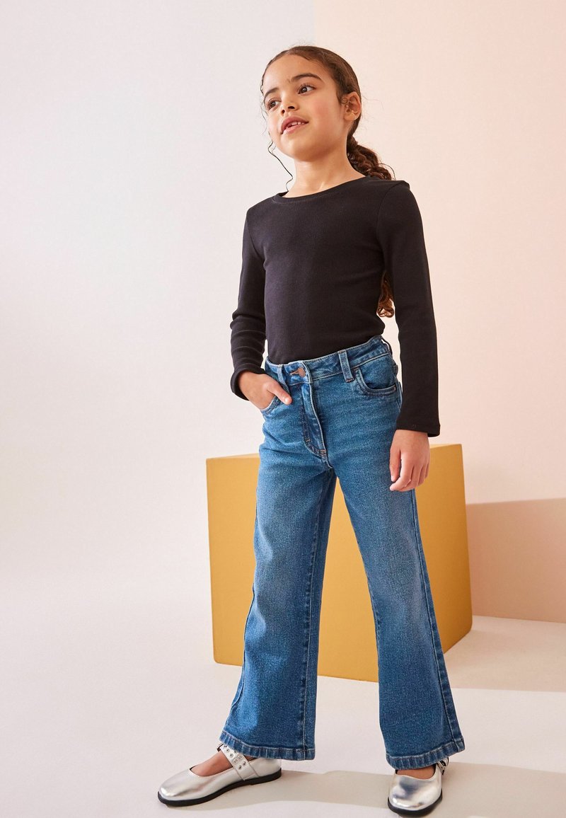 Haut noir à manches longues et jean taille haute évasé bleu clair. Chaussures plates argentées avec des détails de sangle. Fond simple avec un bloc jaune.