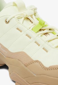 Zapatilla amarilla clara con parte superior de tela texturizada, acentos verdes y suela de goma color canela. Cuenta con cordones elásticos y una lengüeta para ponerla y quitarla fácilmente.