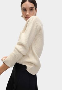 Maglione a costine color crema con maniche lunghe, caratterizzato da una vestibilità rilassata e spacco laterale. Indossato con un completo nero, evidenziando il contrasto testurale.