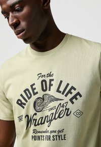 Jasnozielona bawełniana koszulka z czarnym nadrukiem graficznym oraz wzorem skrzydlatych kół z napisem "Ride of Life" i brandingiem "Wrangler".