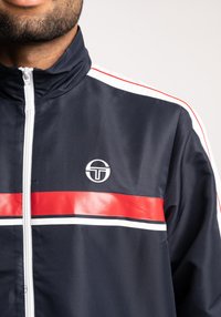 Giacca zip-up blu navy con strisce orizzontali rosse e bianche. Realizzata in materiale liscio e leggero. Presenta un logo circolare sul petto.