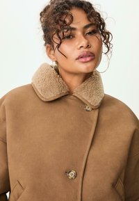 Hnědý kabát z umělé kůže s měkkým béžovým límcem, zapínáním na knoflíky a bočními kapsami, který se vyznačuje jemnou texturou a volným střihem.