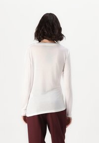 White long-sleeve shirt in lightweight, semi-sheer fabric. Features a round neckline and relaxed fit, paired with maroon trousers.