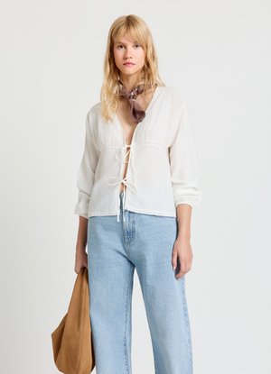 Weiße, leichte Bluse mit Bindeverschluss vorne und Spitzensaum, kombiniert mit hoch taillierten, hellblauen Jeans. Wird mit einer beigefarbenen Tasche geliefert.