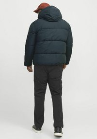 Mørkegrøn dunjakke med hætte, med vandret quiltet design. Bæres med sorte cargo bukser og mørke sneakers.