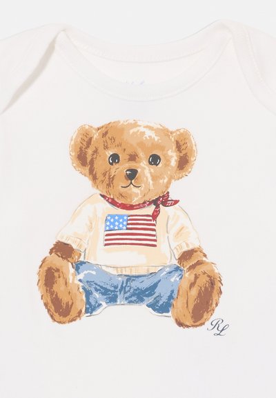 Γραφικό αρκουδάκι σε κρέμα ύφασμα, με καφέ γούνα, μπλε τζιν και κρέμα πουλόβερ με σχέδιο αμερικανικής σημαίας. Έντονα κόκκινο μαντίλι ως αξεσουάρ.