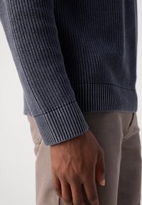 Marineblå ribbestrikket genser med teksturert stoff, med smale mansjetter og subtil horisontal mønster, brukt sammen med lysebeige bukser.