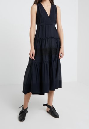 Kvinde iført en sort midi-kjole uden ærmer med dyb V-hals og lagdelt nederdel, kombineret med sorte flade sko med ankelrem.