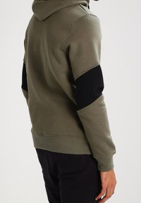 Felpa verde con cappuccio e dettagli neri sulle maniche, realizzata in tessuto morbido, con tasca a marsupio frontale e polsini a costine.