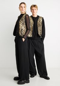 Arany és fekete színű tigrismintás mellények fekete hosszú ujjú felső fölött és bőszárú nadrággal, vastag fekete cipőkkel párosítva.