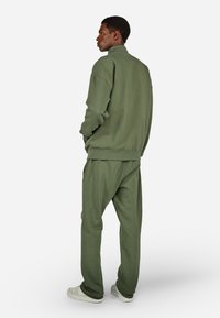 Olīvu zaļa treniņtērps, kurā ietilpst augsta apkakle džemperis un saderīgas bikses ar sānu kabatām. Mīksts audums un brīvs piegriezums. Balti sporta apavi.