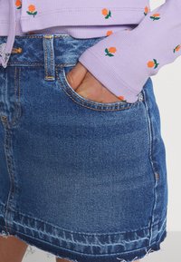 Denim minikjol med fransad fåll, blå färg och orange blommönsterbroderi på en lila långärmad topp. Exponerad sömnad och fickdetaljer synliga.