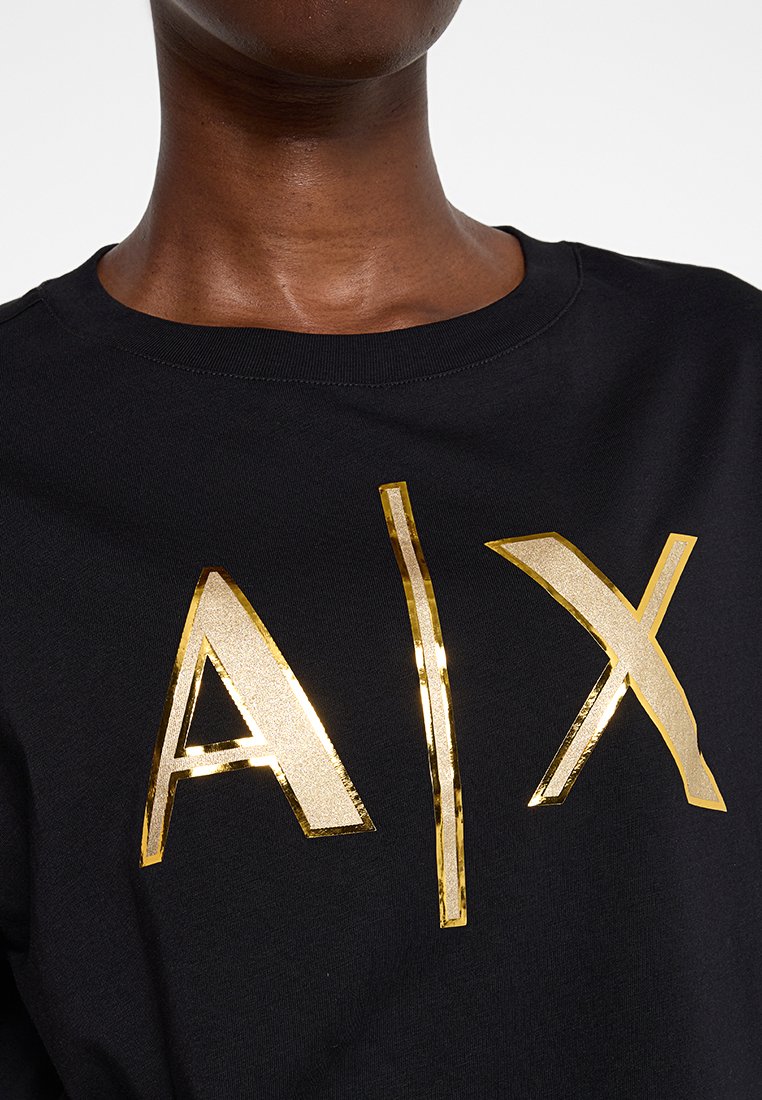 Persona che indossa un maglione nero a girocollo con grandi lettere dorate riflettenti "A|X" sul petto.