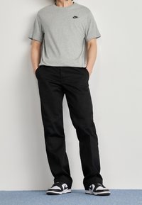 Osoba stojící s rukama v kapsách, oblečená v šedém tričku Nike, černých kalhotách a černobílých teniskách na modrém kobercovém podlaze.