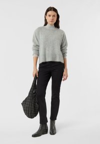 Žena v šedém chlupatém svetru, světlém šedém roláku, černých kalhotách a černých botách, která drží velkou černou prošívanou tašku na neutrálním pozadí.