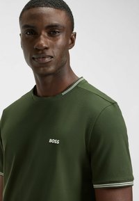 Olive green t-shirt with short sleeves, featuring a contrast striped collar and embroidered white "BOSS" logo on the chest. Smooth texture.