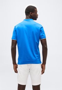 Homme portant un polo bleu vif à manches courtes et un short blanc, debout de dos devant un fond clair uni.